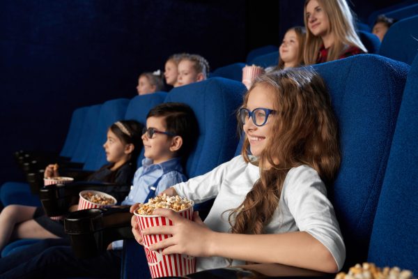 Happy, joyful kids sitting in dark blue, comfortable chairs in modern cinema theatre, watching movie or cartoon. Children enjoying film, smiling, holding buckets with popcorn.